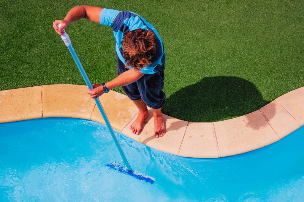 Man stands on the edge of the pool vacuuming the sides of the pool to prevent algae.