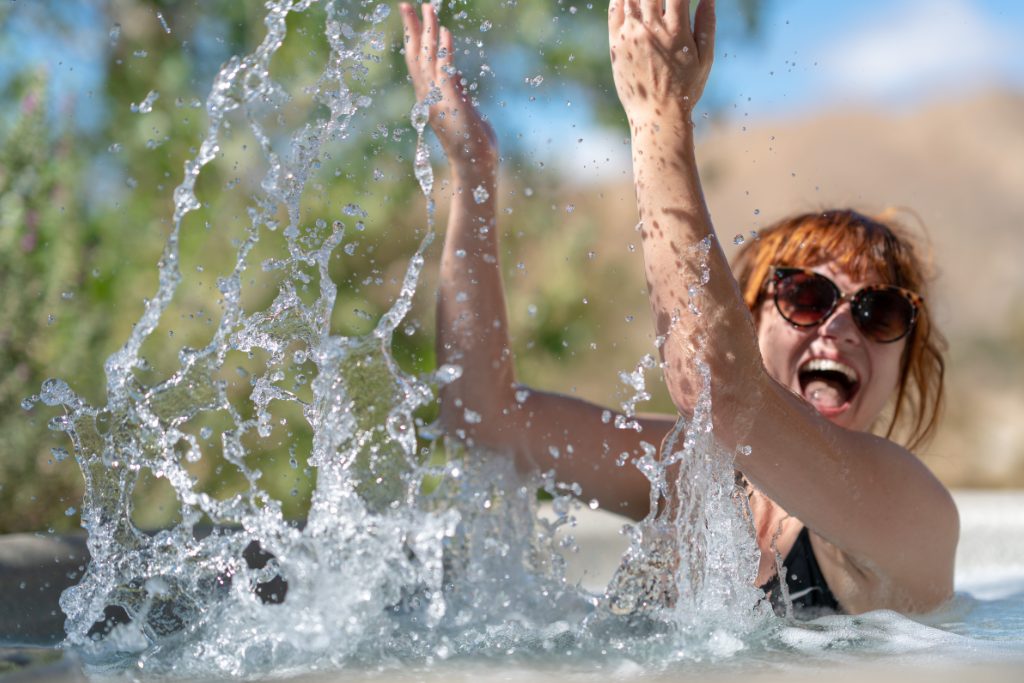 A woman in a black swimsuit and sunglasses splashes water in the hot tub with her hands. She is smiling.