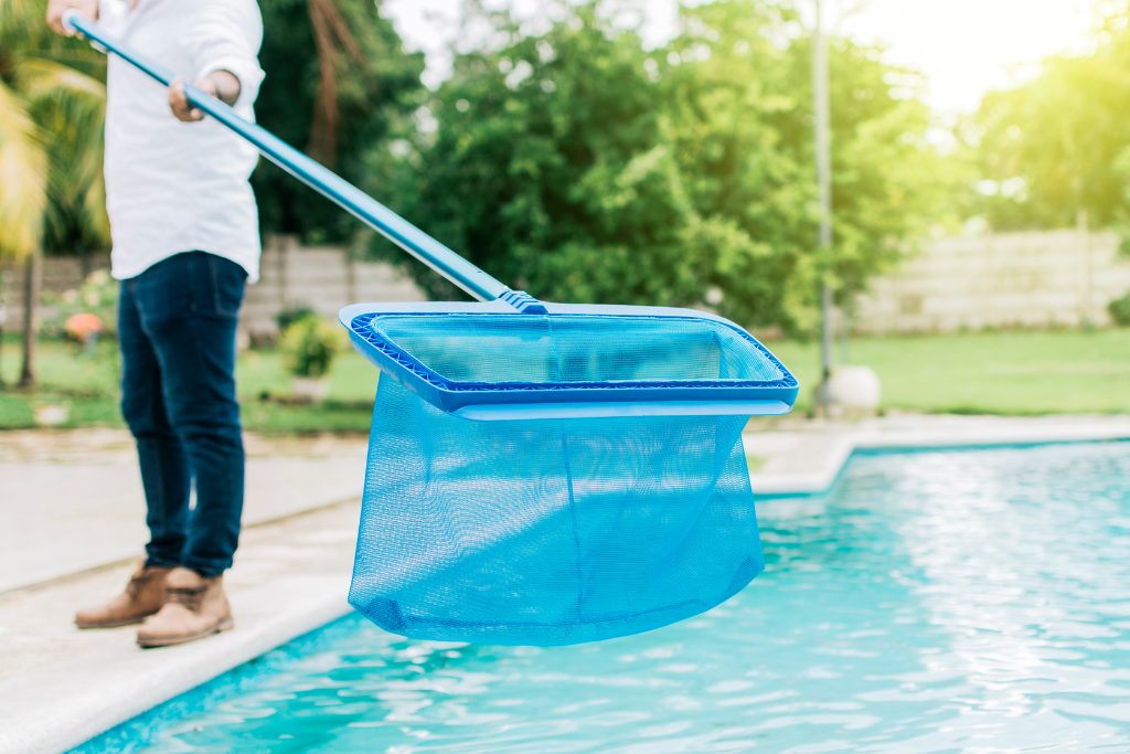 A man in dark pants and white shirt using a blur skimmer to remove debris from an outdoor pool.