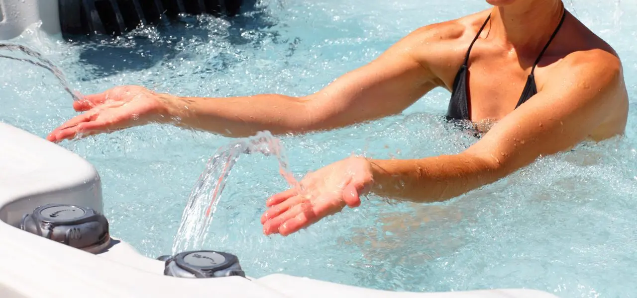 A woman in a black swimsuit reaches forward to feel the hot tub water coming out of the waterfall feature.
