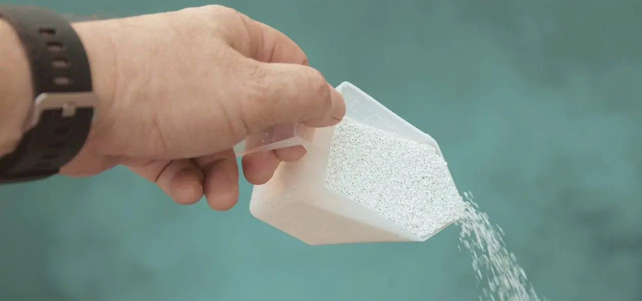 A man's hand dumping chlorine shock into a green tinted pool.