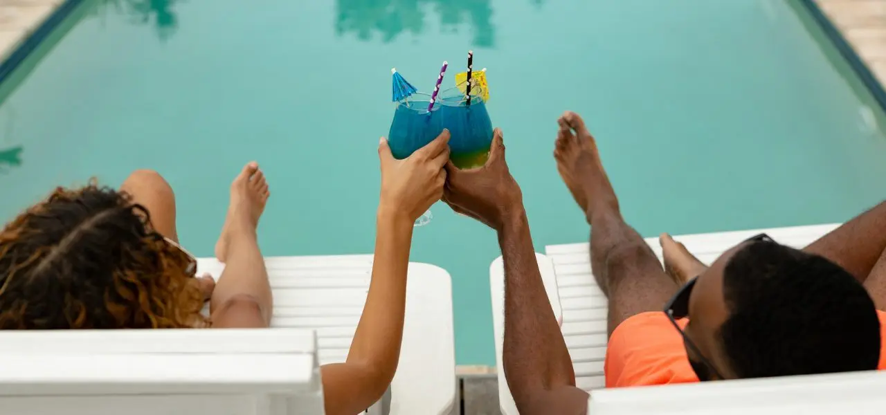 Couple sits on chaise loungers next to each other clinking their drinks. They overlook a clear pool