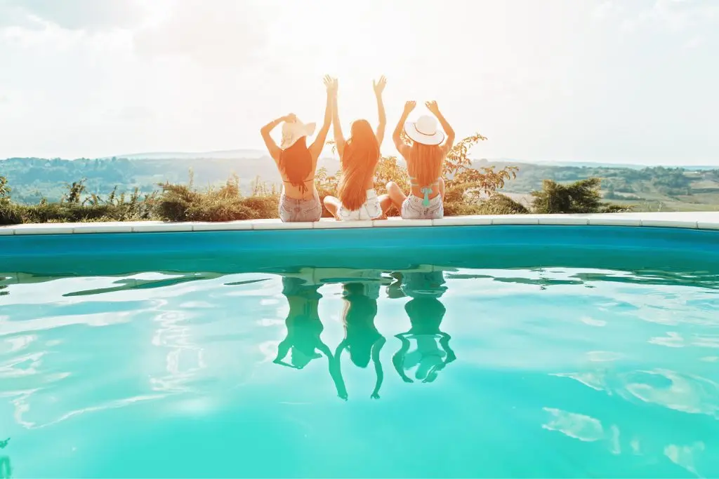 Three women sit on the edge of a pool. Their arms are up in exclamation!