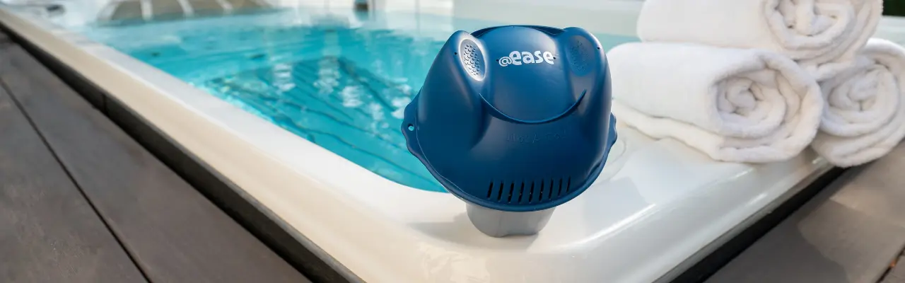 A FROG @ease Sanitizing System for Swim Spas sits on the edge of an outdoor swim spa next to rolled up white towels.