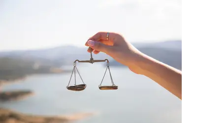 A hand holds a balanced scale. The background is filled with water and mountains.